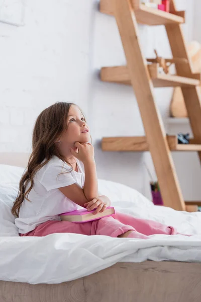 Pensive girl in pajamas looking up while sitting on bed with book — Stock Photo