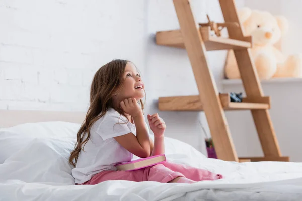 Dreamy girl in pajamas looking up while sitting on bed with book — Stock Photo