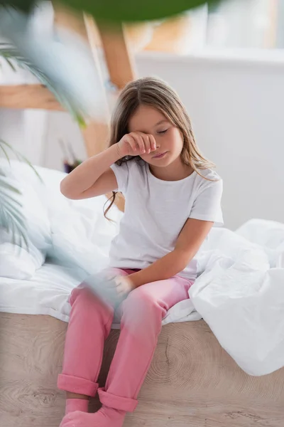 Selective focus of awakened girl in pajamas touching eye while sitting on bed — Stock Photo