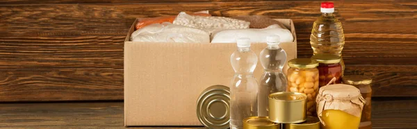 Panoramic shot of cardboard box with groats near water, oil, canned food and honey on wooden background, charity concept — Stock Photo