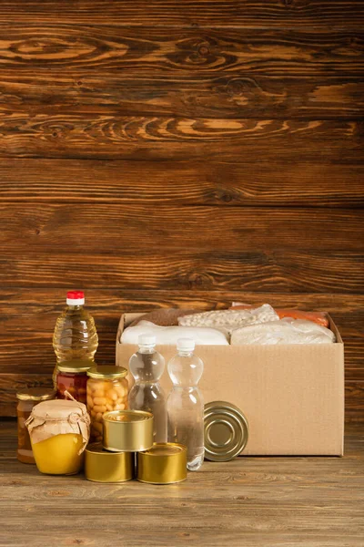 Картонна коробка з крупою біля води, олії, консервованої їжі та меду на дерев'яному фоні, благодійна концепція — стокове фото