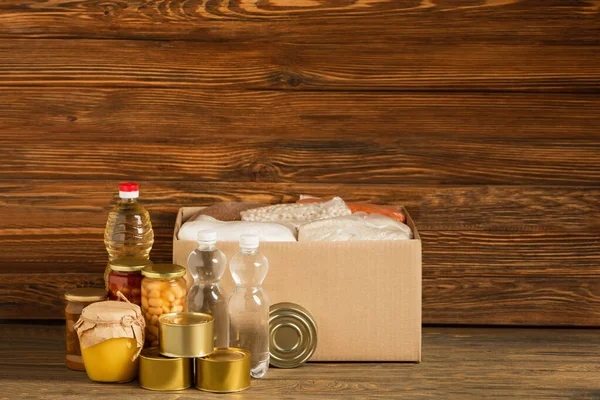 Caja de cartón con grañones cerca del agua, aceite, alimentos enlatados y miel sobre fondo de madera, concepto de caridad - foto de stock