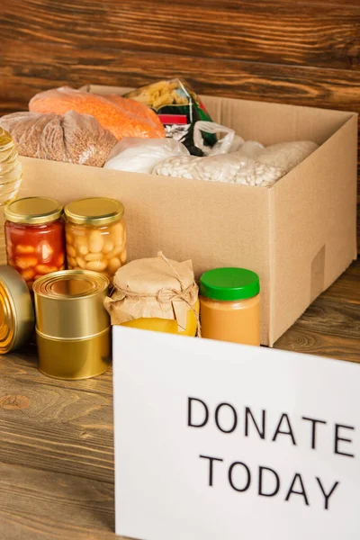 Selective focus of cardboard box with food and donate today card on wooden background, charity concept — Stock Photo