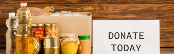 Panoramic shot of cardboard box with groats near water, oil, canned food, honey and donate today card on wooden background, charity concept — Stock Photo