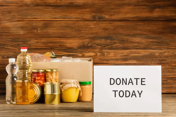 Cardboard box with groats near water, oil, canned food, honey and donate today card on wooden background, charity concept — Stock Photo