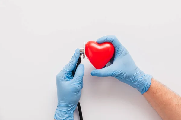 Vista recortada del médico en guantes de látex examinando el corazón rojo con estetoscopio sobre fondo blanco - foto de stock