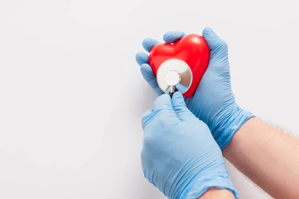 Vista recortada del médico en guantes de látex examinando el corazón rojo con estetoscopio sobre fondo blanco - foto de stock