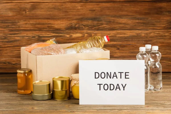 Cardboard box with groats near water, oil, canned food, honey and donate today card on wooden background, charity concept — Stock Photo