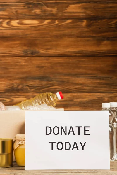 Cardboard box with food and donate today card on wooden background, charity concept — Stock Photo
