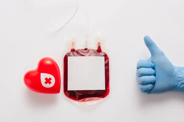 Cropped view of doctor showing thumb up near blood package and red heart on white background — Stock Photo