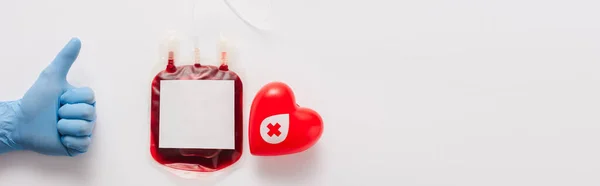 Vista cortada do médico mostrando polegar para cima perto de pacote de sangue e coração vermelho no fundo branco, tiro panorâmico — Fotografia de Stock