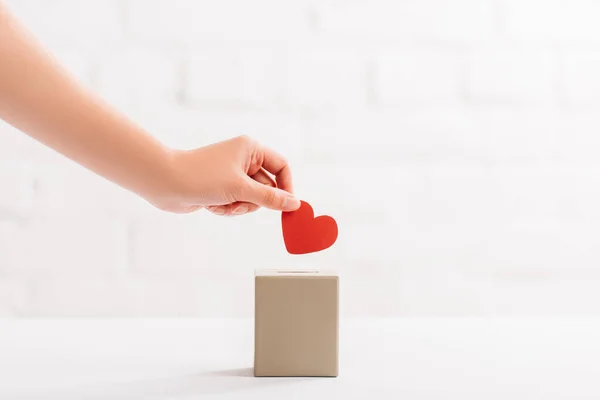 Vista recortada de la mano femenina poniendo corazón rojo en caja sobre fondo blanco, concepto de donación - foto de stock