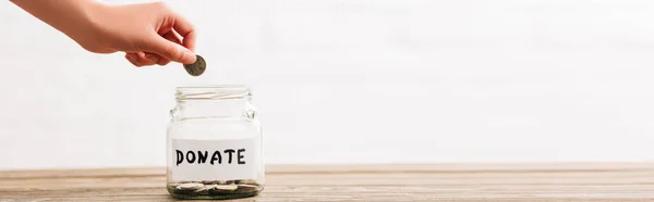 Cropped view of woman putting coin in penny jar with donate lettering on wooden surface on white background, panoramic shot — Stock Photo