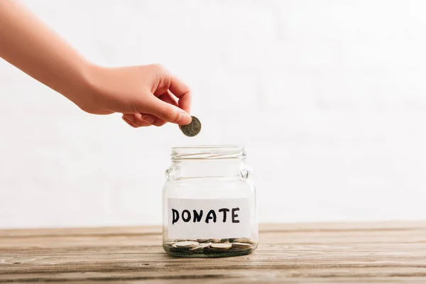 Cropped view of woman putting coin in penny jar with donate lettering on wooden surface on white background — Stock Photo