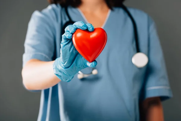Обрізаний вид на лікаря зі стереоскопом в латексній рукавичці, що тримає червоне серце ізольовано на сірому — стокове фото