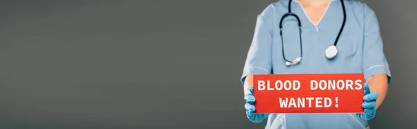 Cropped view of doctor with stereoscope holding red card with blood donors wanted lettering isolated on grey, panoramic shot — Stock Photo