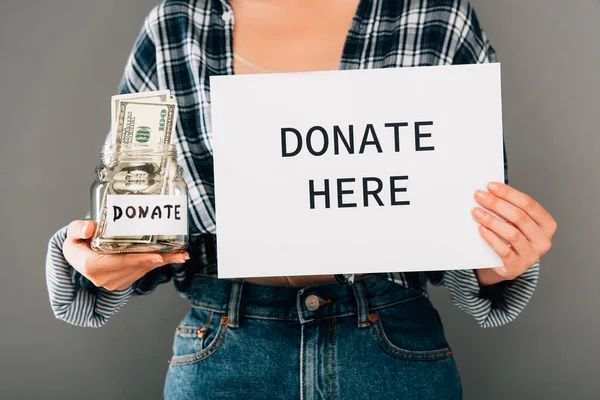 Cropped view of woman holding card with donate here lettering and jar with money on grey background — Stock Photo