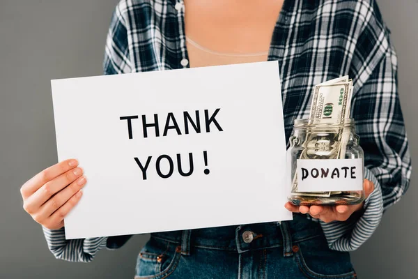 Cropped view of woman holding card with thank you lettering and jar with donated money on grey background — Stock Photo