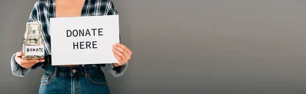 Cropped view of woman holding card with donate here lettering and jar with money on grey background, panoramic shot — Stock Photo