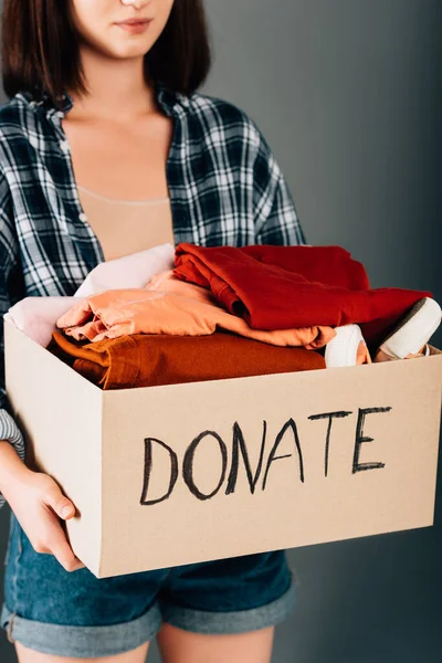 Cropped view of woman holding box with donate lettering and clothes on grey background — Stock Photo