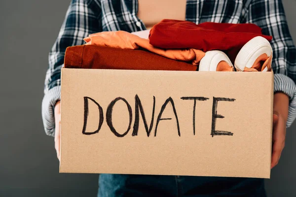 Cropped view of woman holding box with donate lettering and clothes on grey background — Stock Photo