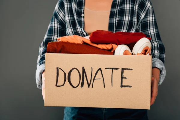 Cropped view of woman holding box with donate lettering and clothes on grey background — Stock Photo