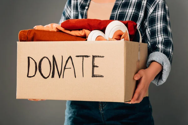Cropped view of woman holding box with donate lettering and clothes on grey background — Stock Photo