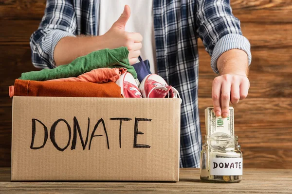 Abgeschnittene Ansicht eines Mannes, der den Daumen in der Nähe eines Kartons mit gespendeten Kleidungsstücken und Geld auf Holzgrund zeigt, Charity-Konzept — Stockfoto