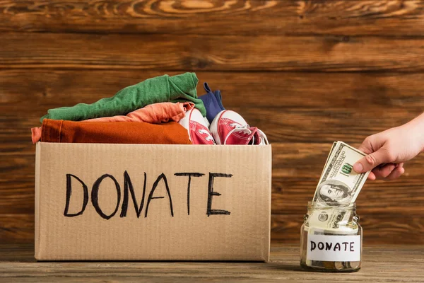 Vista recortada del hombre poniendo dinero en frasco cerca de caja de cartón con donar letras y ropa sobre fondo de madera, concepto de caridad - foto de stock