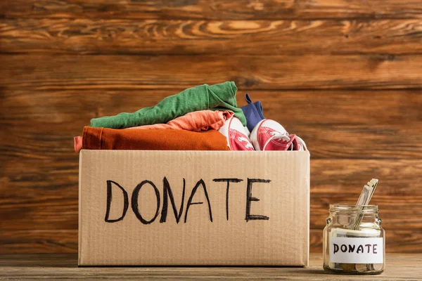 Argent dans un bocal près de la boîte en carton avec des dons de lettres et de vêtements sur fond en bois, concept de charité — Stock Photo