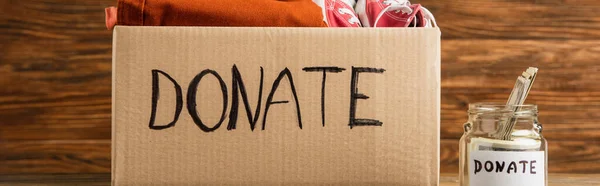 Panoramic shot of money in jar near cardboard box with donate lettering and clothes on wooden background, charity concept — Stock Photo