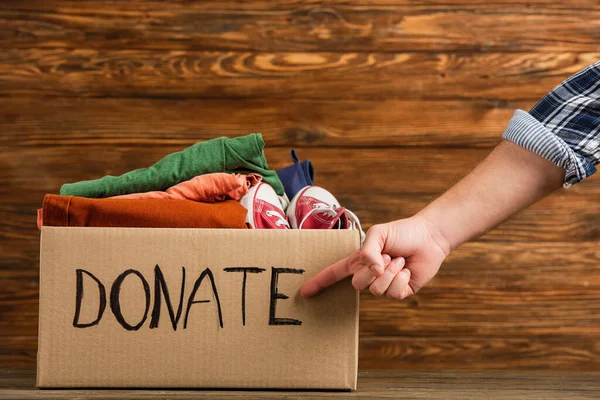 Cropped view of man pointing at cardboard box with donate lettering and clothes on wooden background, charity concept — Stock Photo