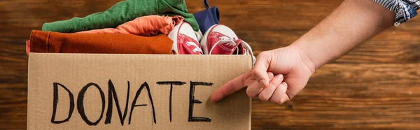 Cropped view of man pointing at cardboard box with donate lettering and clothes on wooden background, charity concept — Stock Photo