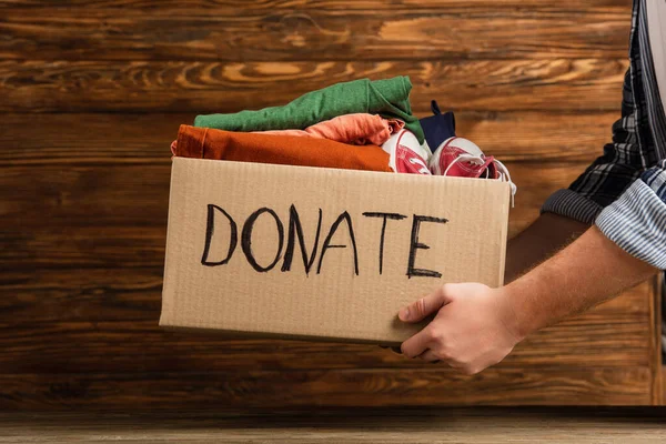 Cropped view of man holding cardboard box with donate lettering and clothes on wooden background, charity concept — Stock Photo