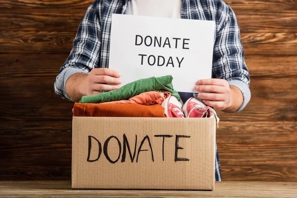 Vista recortada del hombre sosteniendo donar hoy tarjeta cerca de la caja de cartón con ropa sobre fondo de madera, concepto de caridad - foto de stock