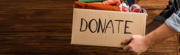 Cropped view of man holding cardboard box with donate lettering and clothes on wooden background, charity concept — Stock Photo