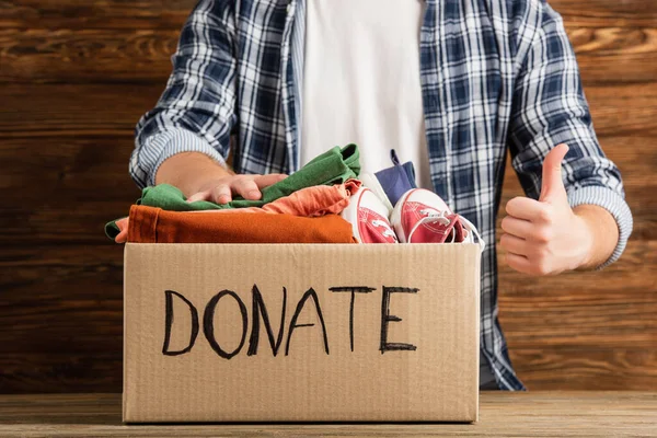 Cropped view of man showing thumb up near cardboard box with donate lettering and clothes on wooden background, charity concept — Stock Photo