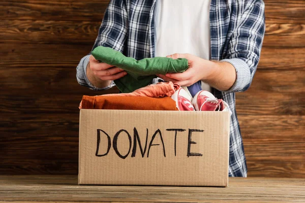 Cropped view of man putting clothes in cardboard box with donate lettering on wooden background, charity concept — Stock Photo