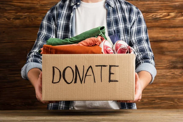 Cropped view of man holding cardboard box with donate lettering and clothes on wooden background, charity concept — Stock Photo
