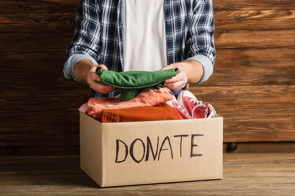 Cropped view of man putting clothes in cardboard box with donate lettering on wooden background, charity concept — Stock Photo