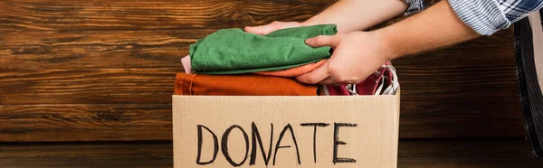 Cropped view of man putting clothes in cardboard box with donate lettering on wooden background, charity concept — Stock Photo