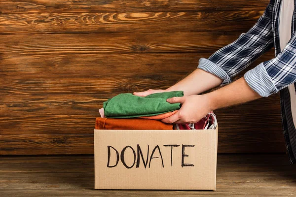 Cropped view of man putting clothes in cardboard box with donate lettering on wooden background, charity concept — Stock Photo
