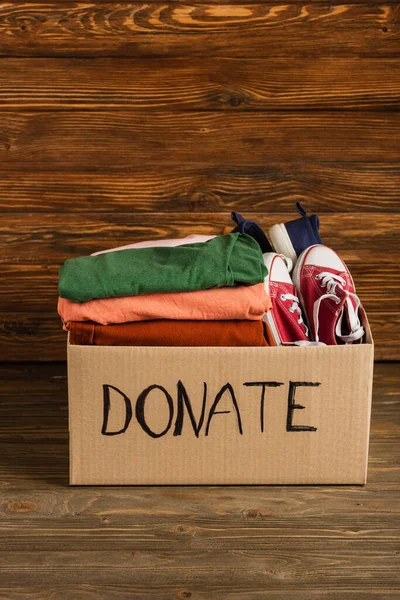 Cardboard box with donated clothes and footwear on wooden background, charity concept — Stock Photo