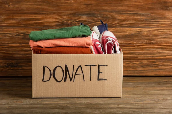 Cardboard box with donated clothes and footwear on wooden background, charity concept — Stock Photo