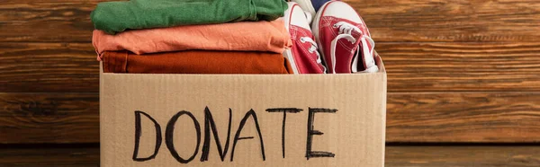 Panoramic shot of cardboard box with donated clothes and footwear on wooden background, charity concept — Stock Photo