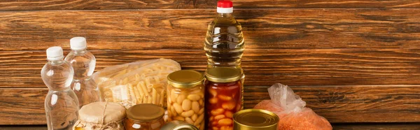 Panoramic shot of groats near water, oil, canned food and honey on wooden background, charity concept — Stock Photo