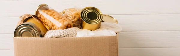 Panoramic shot of cardboard box with donated food on wooden background, charity concept — Stock Photo