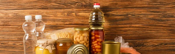 Panoramic shot of groats near water, oil, canned food and honey on wooden background, charity concept — Stock Photo
