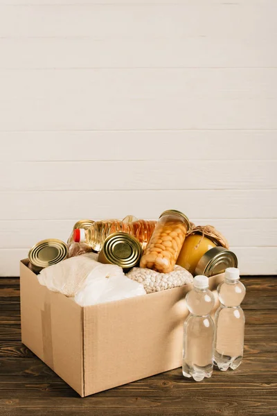 Cardboard box with donated food and water on wooden background, charity concept — Stock Photo