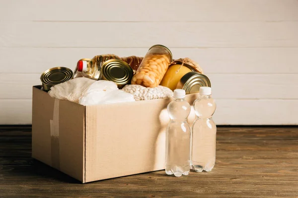 Cardboard box with donated food and water on wooden background, charity concept — Stock Photo